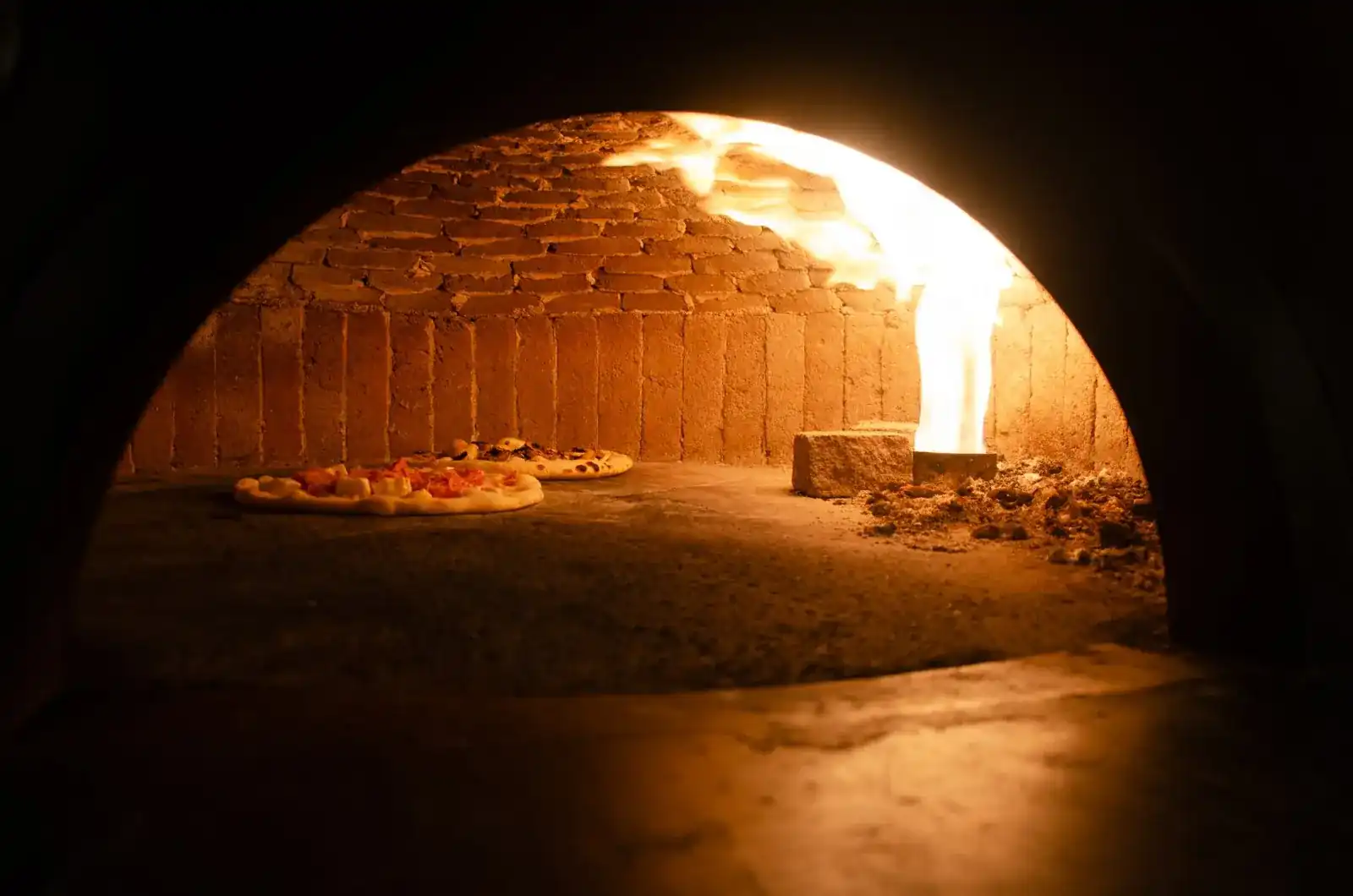 Inside the brick oven: two pizzas on the stone floor and a flame rising from the wood fire