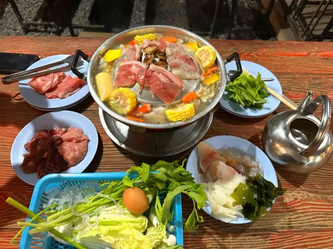 A moo kratha pan with pork on the dome and corn, carrot and pumpkin in the broth, with plates of raw meat and a basket of cabbage, glass noodles and an egg on a wooden table