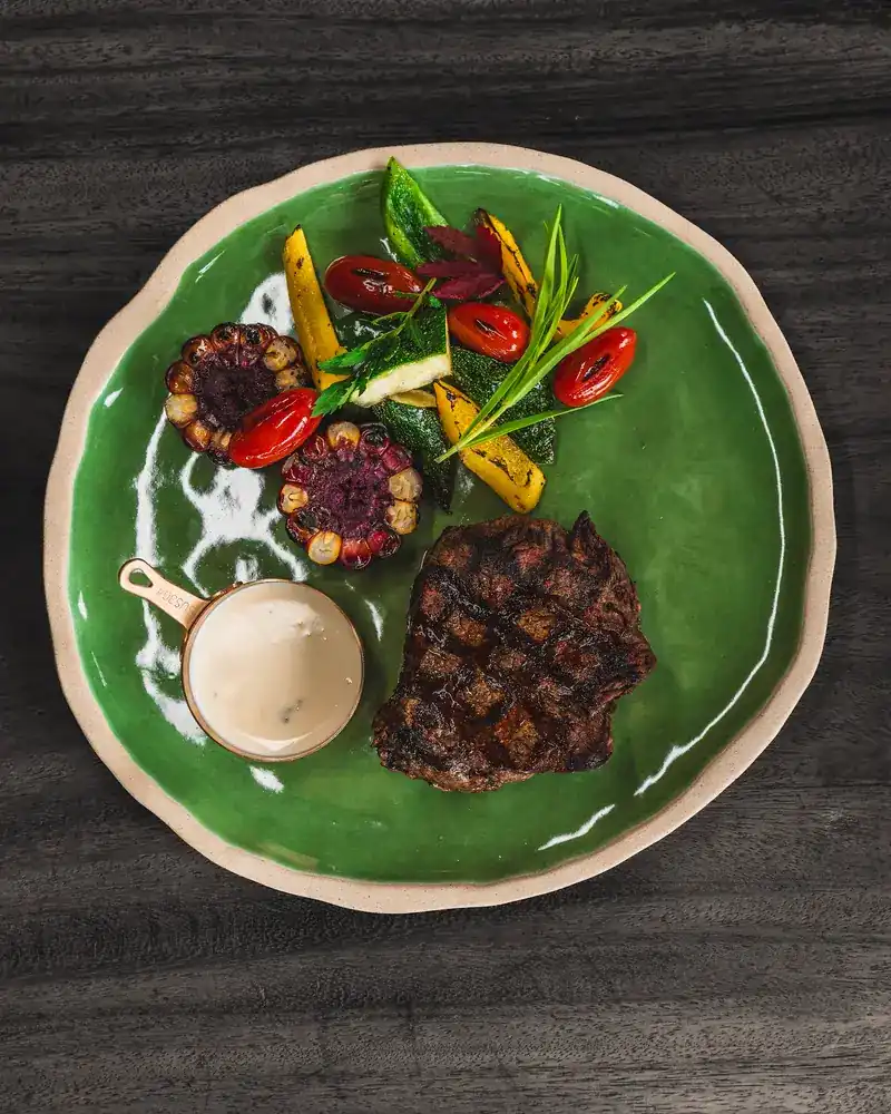 A grilled steak on a green plate with grilled vegetables, cherry tomatoes and a small pot of sauce