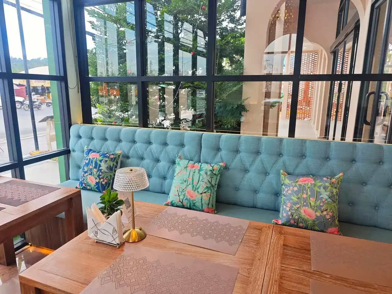 A turquoise button-back sofa with floral cushions and a brass lamp, beside tall black-framed windows