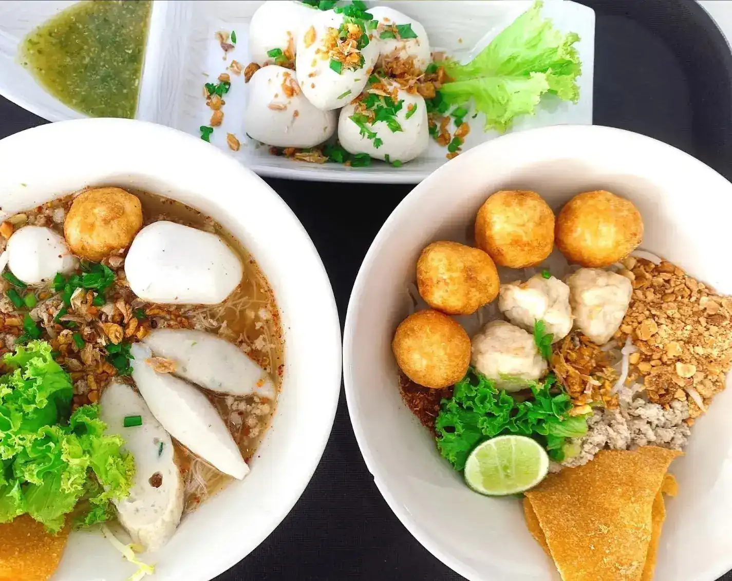 Two bowls of fish ball noodles, one in soup and one dry, with a plate of steamed fish balls and green dipping sauce