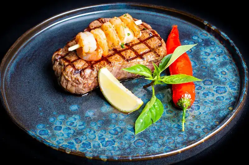 Grilled steak topped with a prawn skewer, with lime and red chili on a blue plate