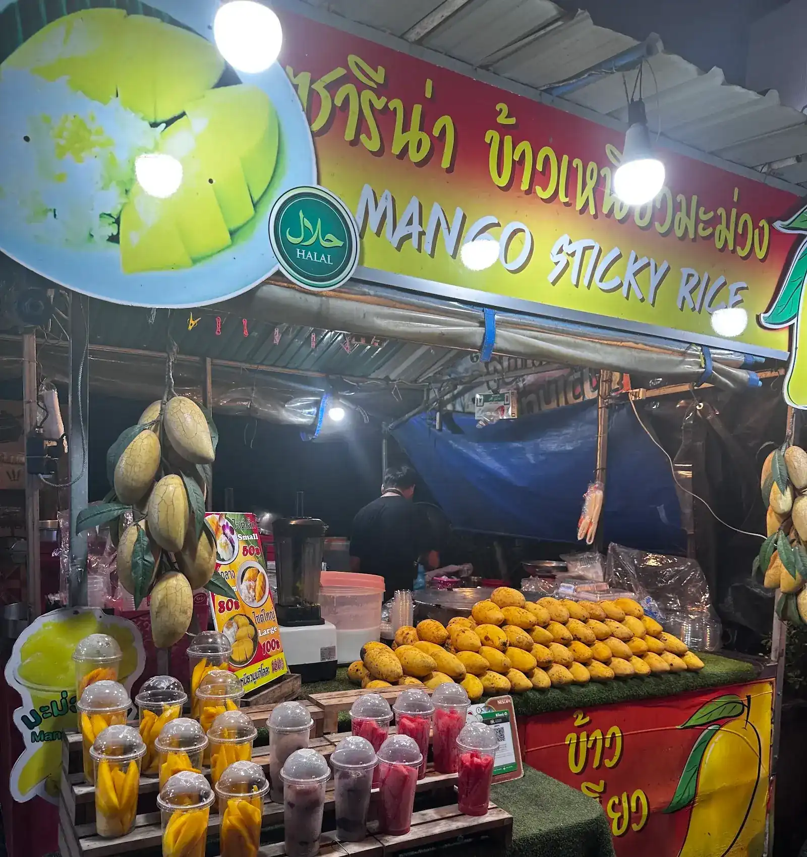 The Sarina stall at night: a yellow and red sign reading Mango Sticky Rice, bunches of mangoes hanging, ripe mangoes piled on the counter and cups of cut fruit in front