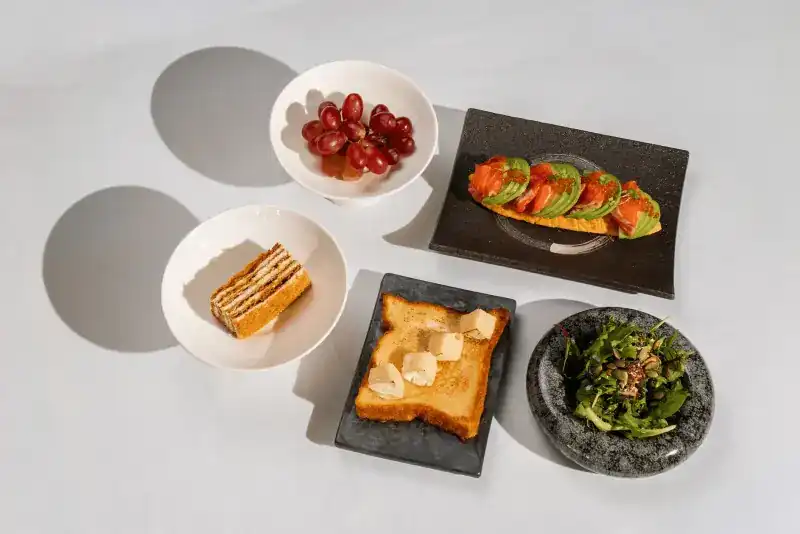 A breakfast set of salmon and avocado toast, toast with cheese, green salad, grapes and a slice of layered cake