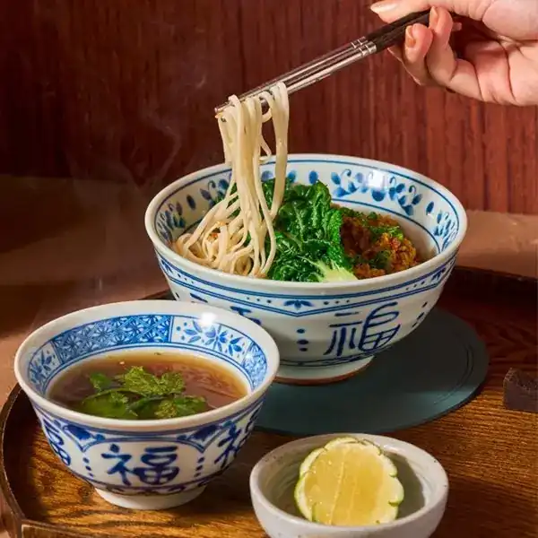 Noodles lifted with chopsticks from a blue and white bowl, with a bowl of broth beside it