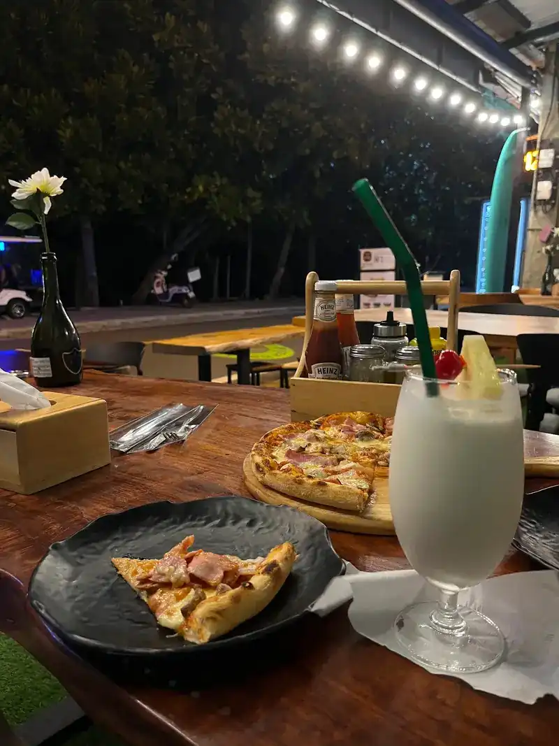 Evening at a wooden table: a pizza and a tall creamy drink with a cherry, lights along the roof