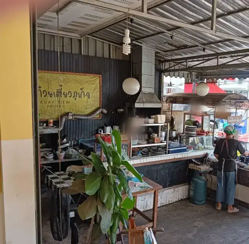The open kitchen and the shop's yellow sign under a tin roof, with Thep Krasattri Road beyond