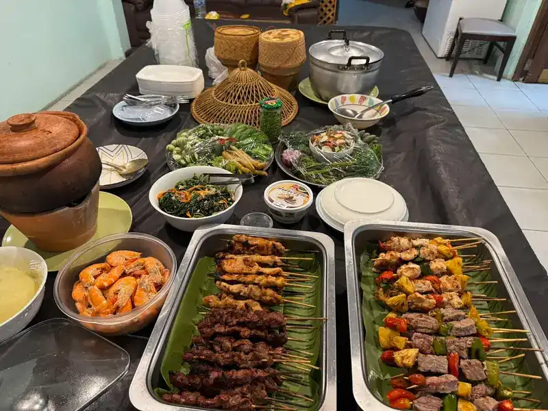 A long table set with skewers, a bowl of prawns, fresh greens and sticky rice baskets
