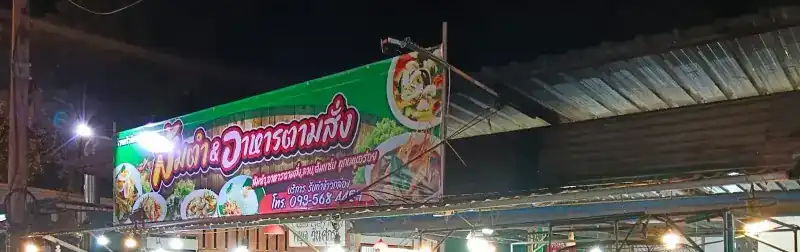 The shop's green sign lit up at night, reading ส้มตำ & อาหารตามสั่ง