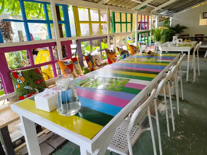 A long table painted in rainbow stripes, with patterned cushions on the bench and coloured window frames behind