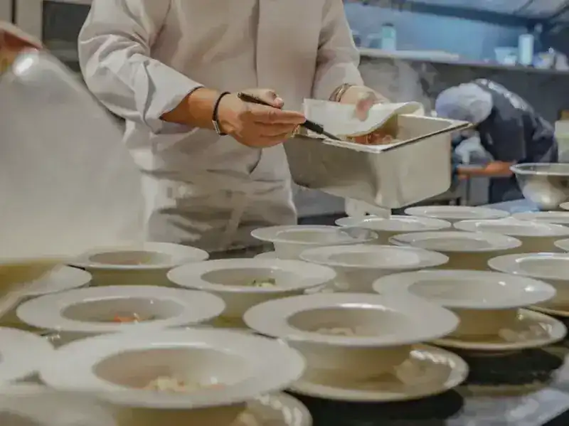 A chef's hands plating a row of white bowls in the SUAY kitchen
