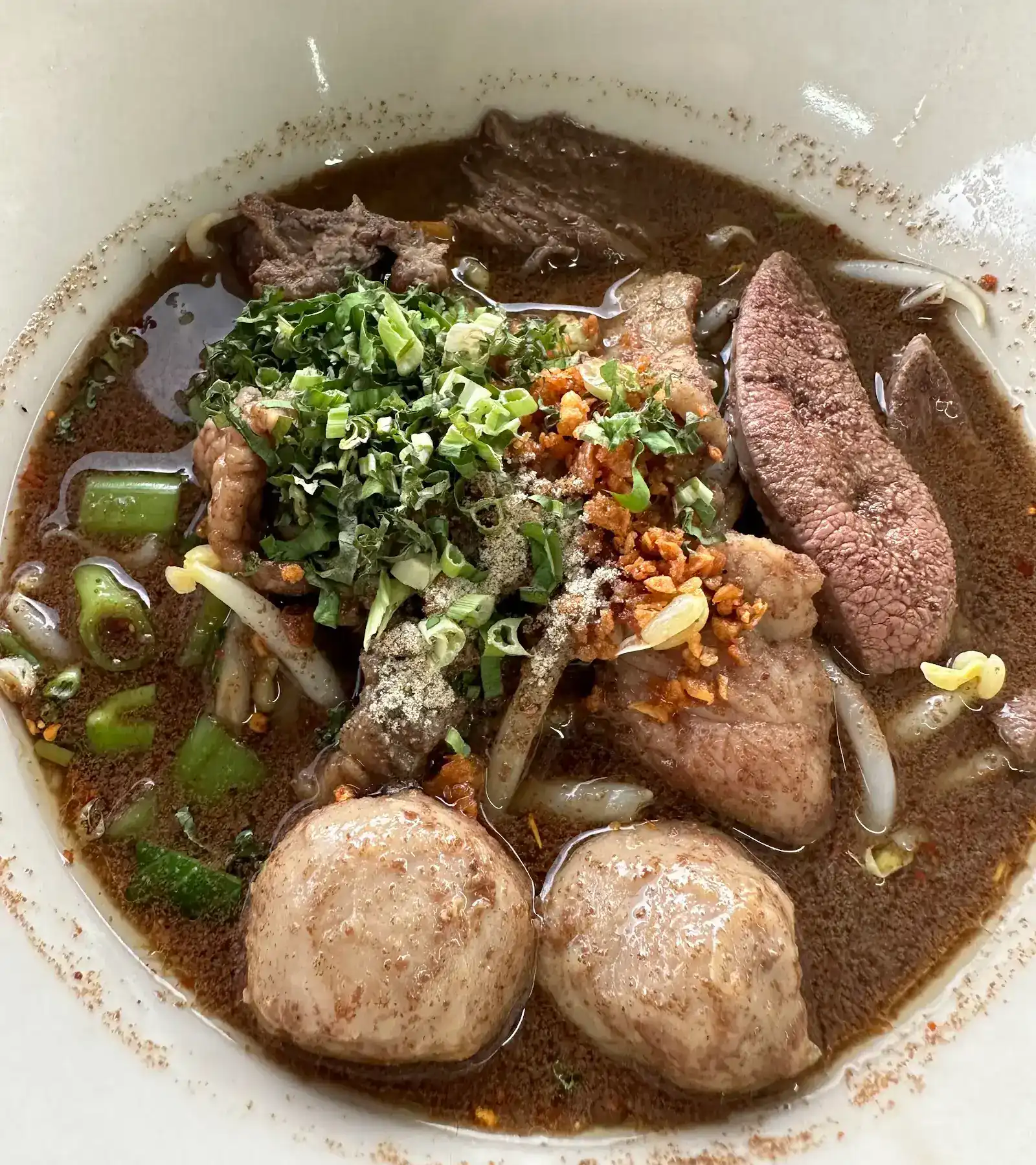A bowl of boat noodles: dark broth, two meatballs, liver, sliced meat, herbs and fried garlic