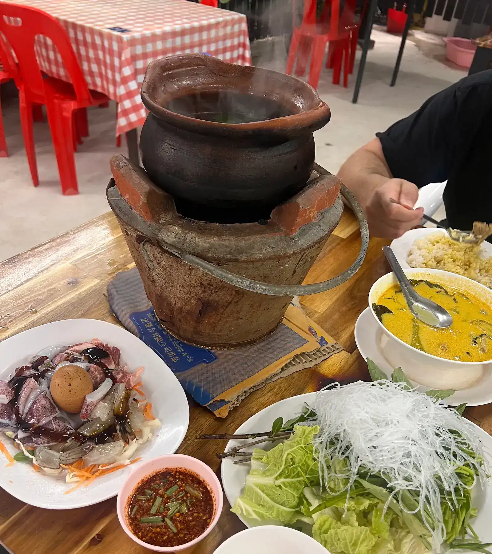 A clay pot steaming on a charcoal stove, with raw seafood, glass noodles, greens, dipping sauce and a curry beside it