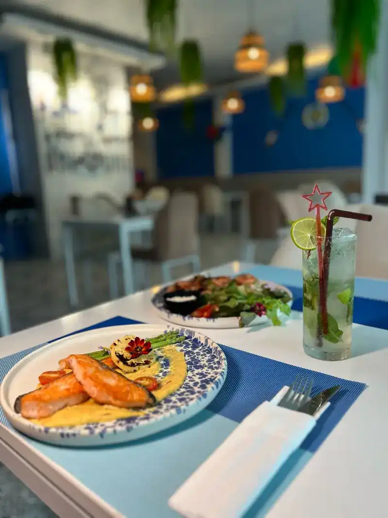 Grilled salmon and a tall glass with lime on a blue placemat, the dining room behind