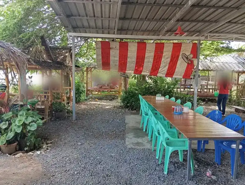 The open dining area: a long wooden table with green chairs under a tin roof and a striped canvas, with sala huts in the garden behind