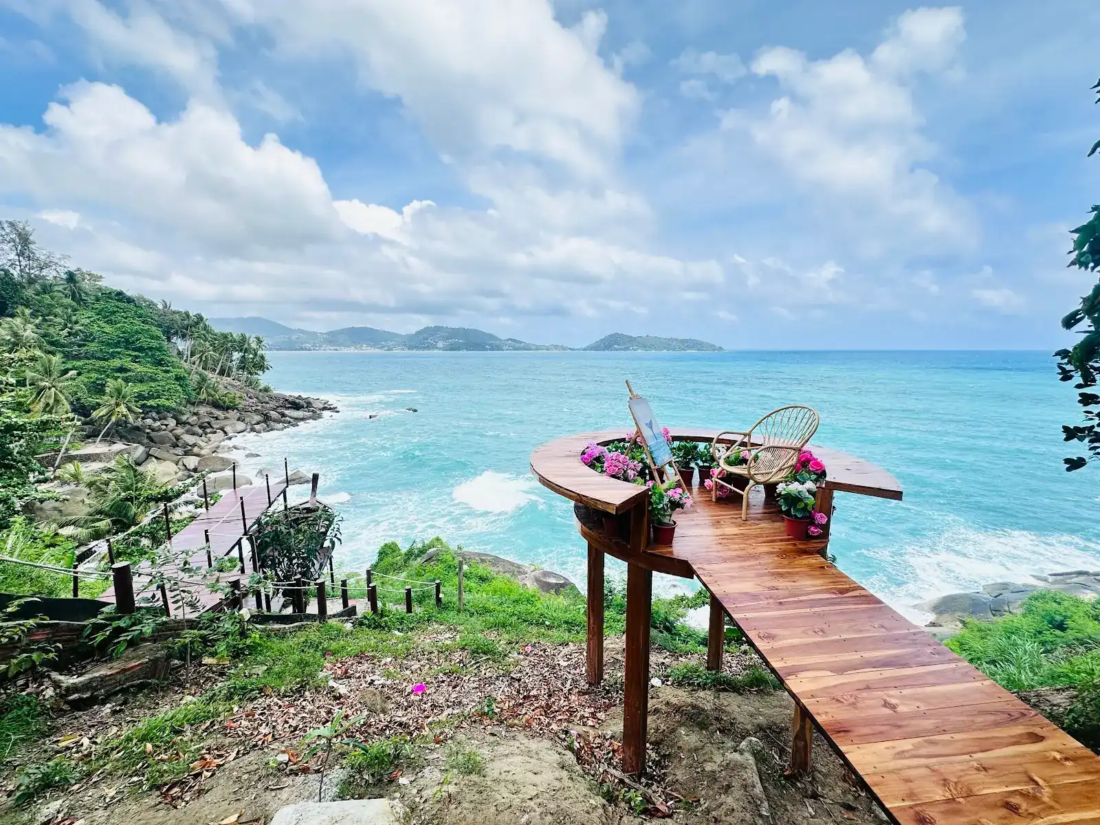 A wooden walkway out to a round platform with a rattan chair and pink flowers, over a turquoise sea / ทางเดินไม้ทอดไปยังลานวงกลม มีเก้าอี้หวายและดอกไม้สีชมพู เหนือทะเลสีฟ้าคราม