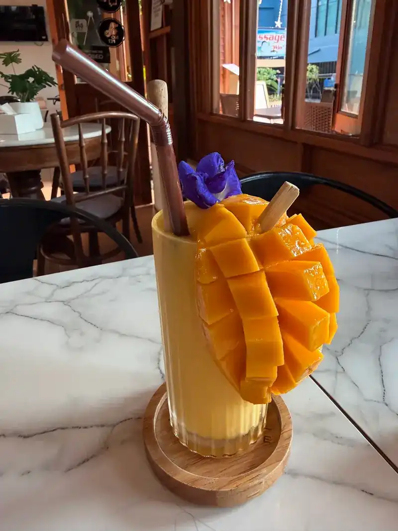 A mango smoothie in a tall glass, topped with cut mango and a butterfly-pea flower
