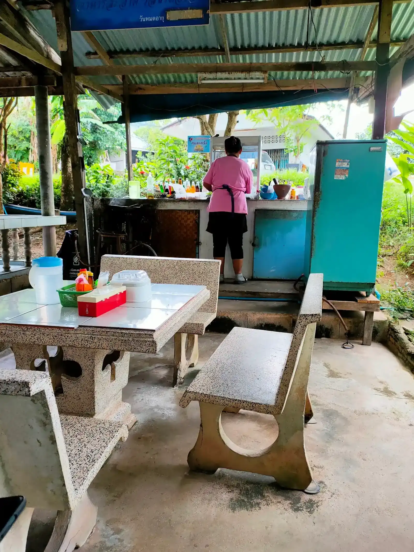 Under the shop's tin roof: stone tables and benches, the cook at the counter with her back to us, plants all around