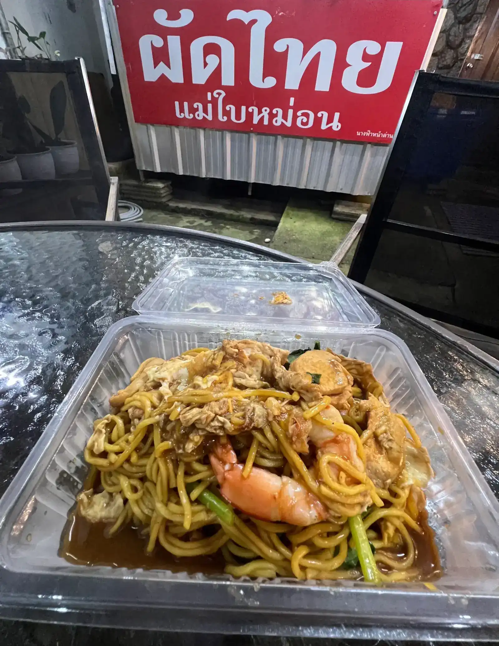 A box of yellow noodles fried with prawns and greens on a glass table, with the red ผัดไทย แม่ใบหม่อน sign behind it
