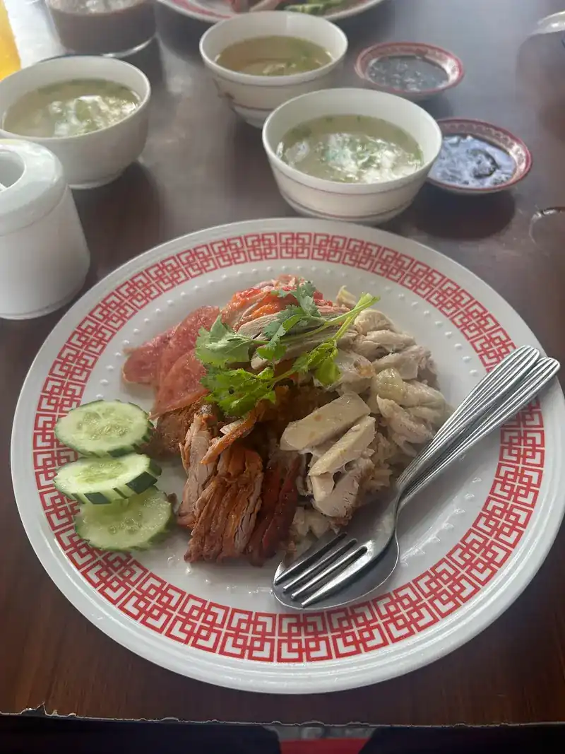 A mixed plate of red pork and chicken on a red-patterned plate, with bowls of soup behind