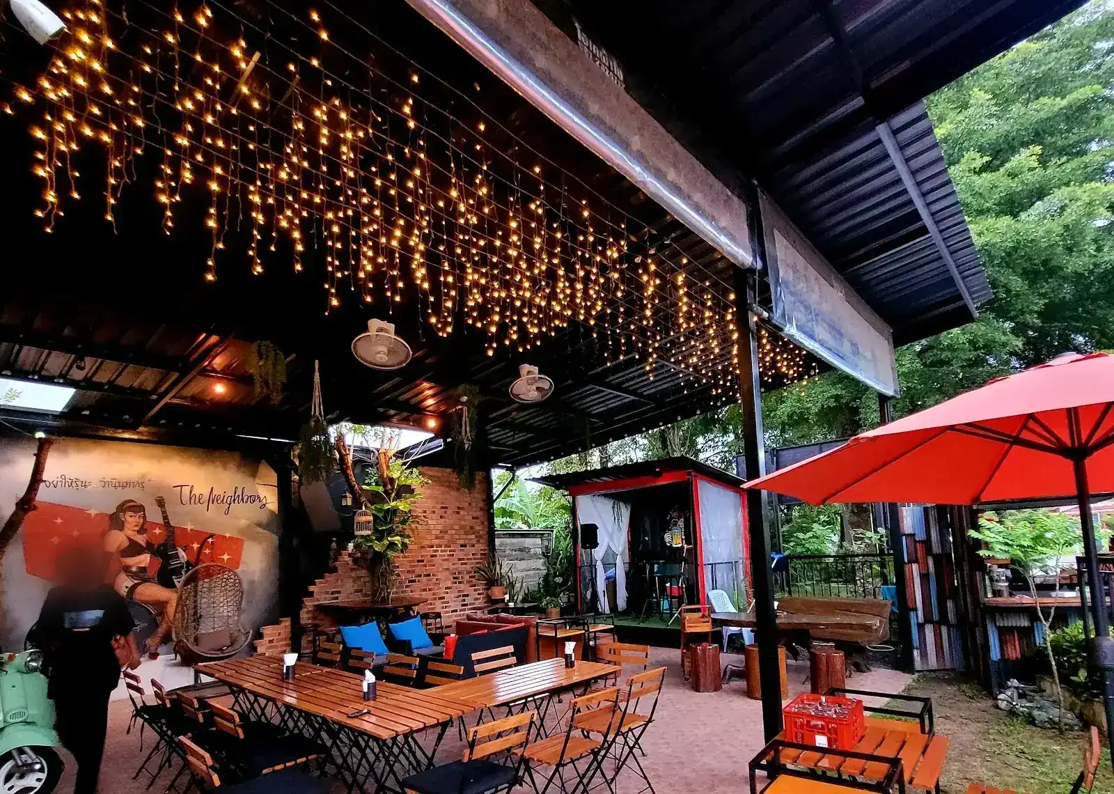 The garden terrace under a canopy of warm string lights, with long wooden tables and a red umbrella