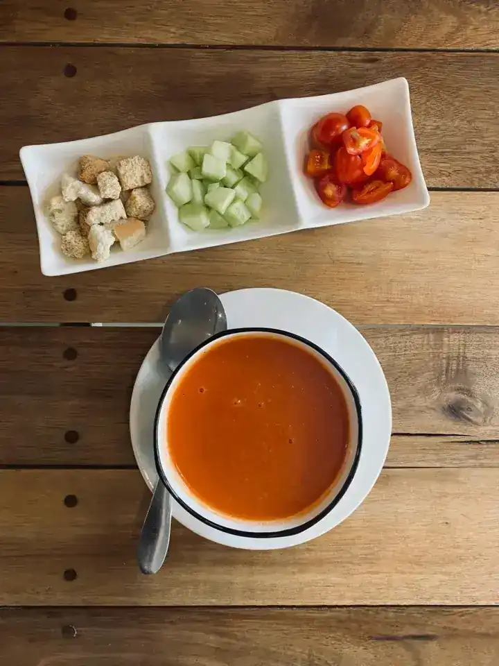 A bowl of gazpacho on a wooden table with croutons, cucumber and tomato on the side