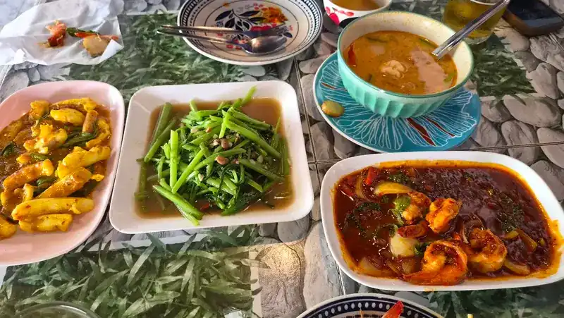 Shared dishes on a pebble-print table: stir-fried morning glory, prawns in a red sauce, a yellow stir-fry and a bowl of soup