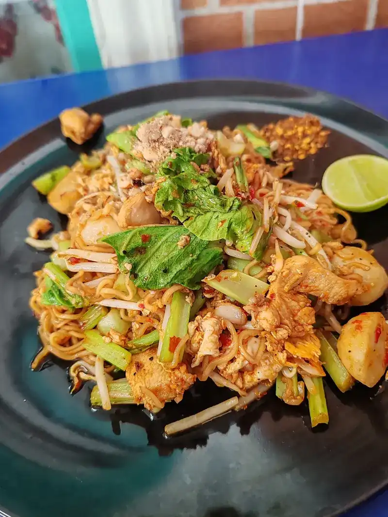 A dark plate of fried noodles with chicken, fish balls, bean sprouts, greens, chilli flakes and lime on a blue table