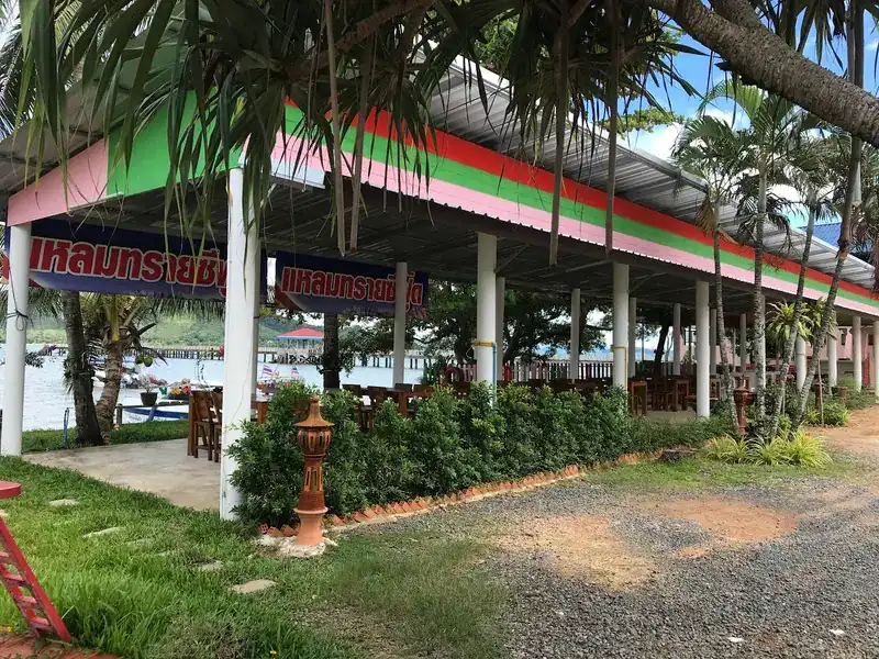 The open dining pavilion with red, green and pink stripes on the roof edge, wooden tables, and the sea and pier behind / ศาลาที่นั่งรับประทานอาหารแบบโล่ง ชายคาทาสีแดง เขียว ชมพู โต๊ะไม้ และทะเลกับสะพานท่าเรือด้านหลัง