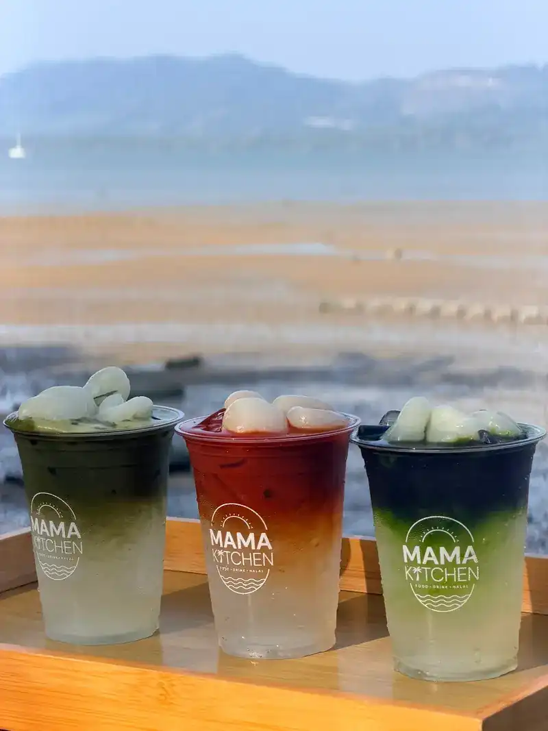 Three layered iced drinks in green, red and blue, in Mama Kitchen cups, with the beach behind