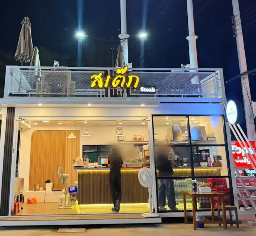 The white glass-walled kitchen at night, with the yellow สเต๊ก sign glowing on its roof rail