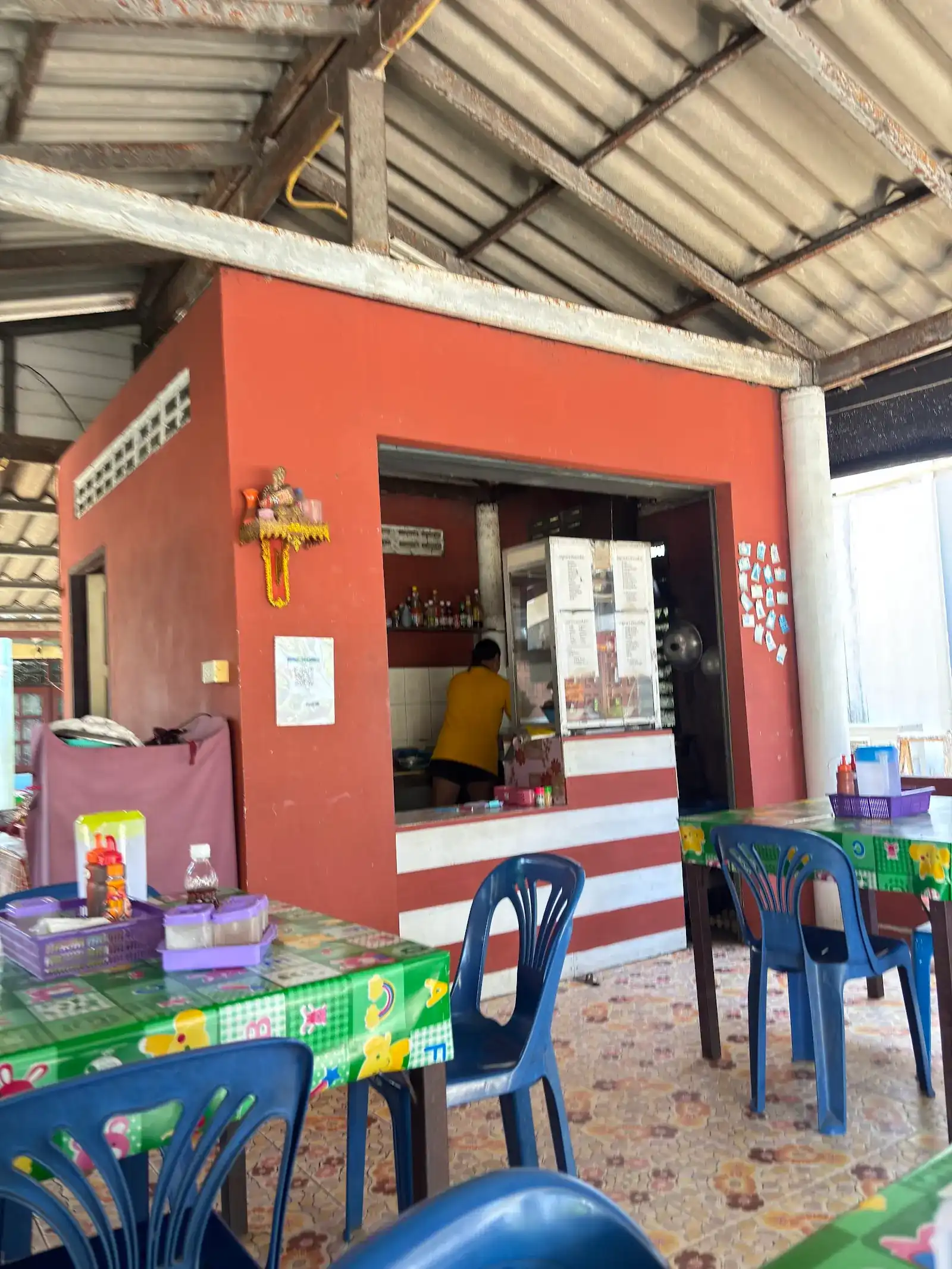 The shop's red kitchen kiosk under a tin roof, with blue plastic chairs and green oilcloth tables; the cook works inside, back turned