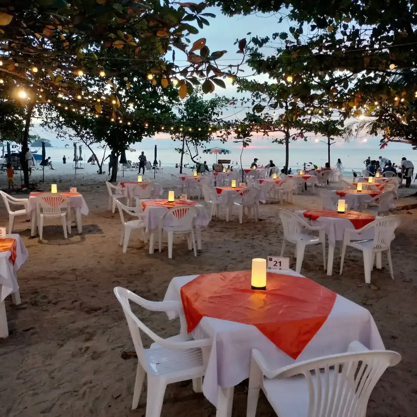 Tables with white cloths, red cloths laid across them and small lamps, set on the sand of Nai Yang Beach at dusk under trees hung with lights