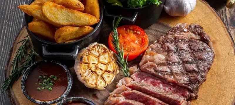Sliced grilled steak on a wooden board with roasted garlic, tomato and potato wedges