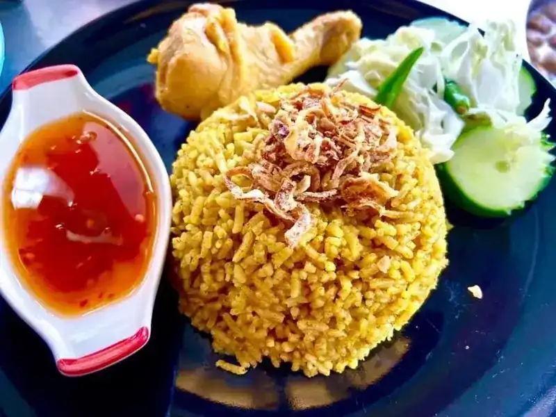Yellow biryani rice topped with fried shallots, a chicken leg, cabbage, cucumber and a dish of red sauce
