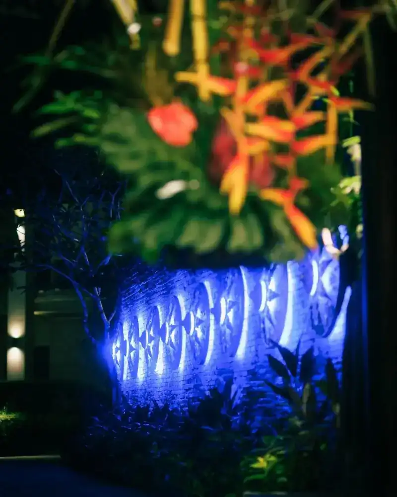 Blue-lit carved shields along a brick wall beneath red and orange tropical flowers