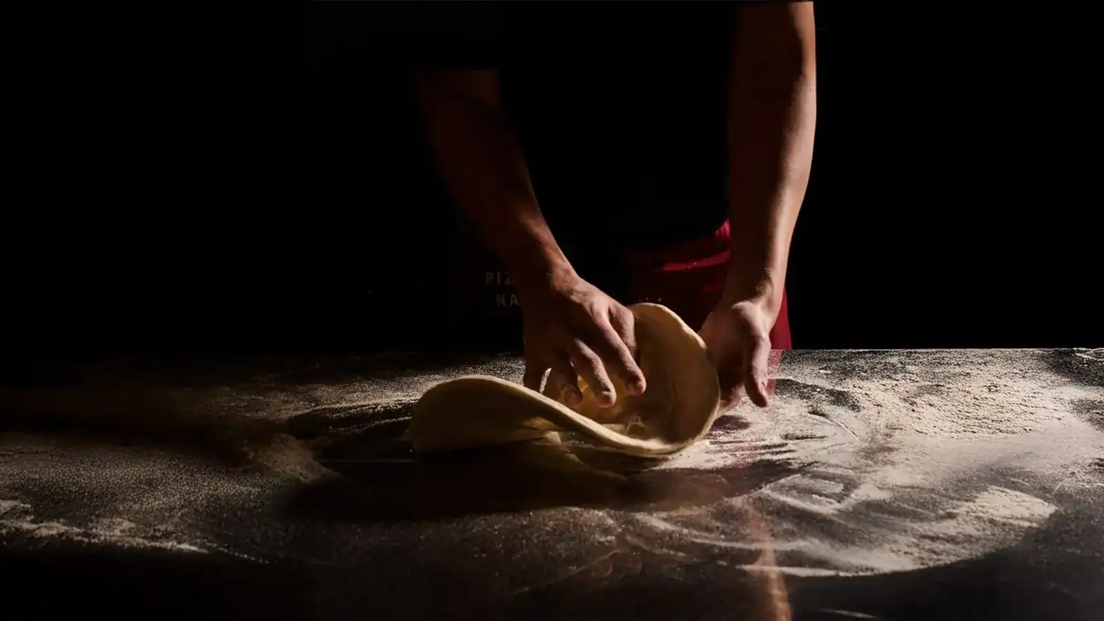 Hands stretching pizza dough on a black, flour-dusted bench