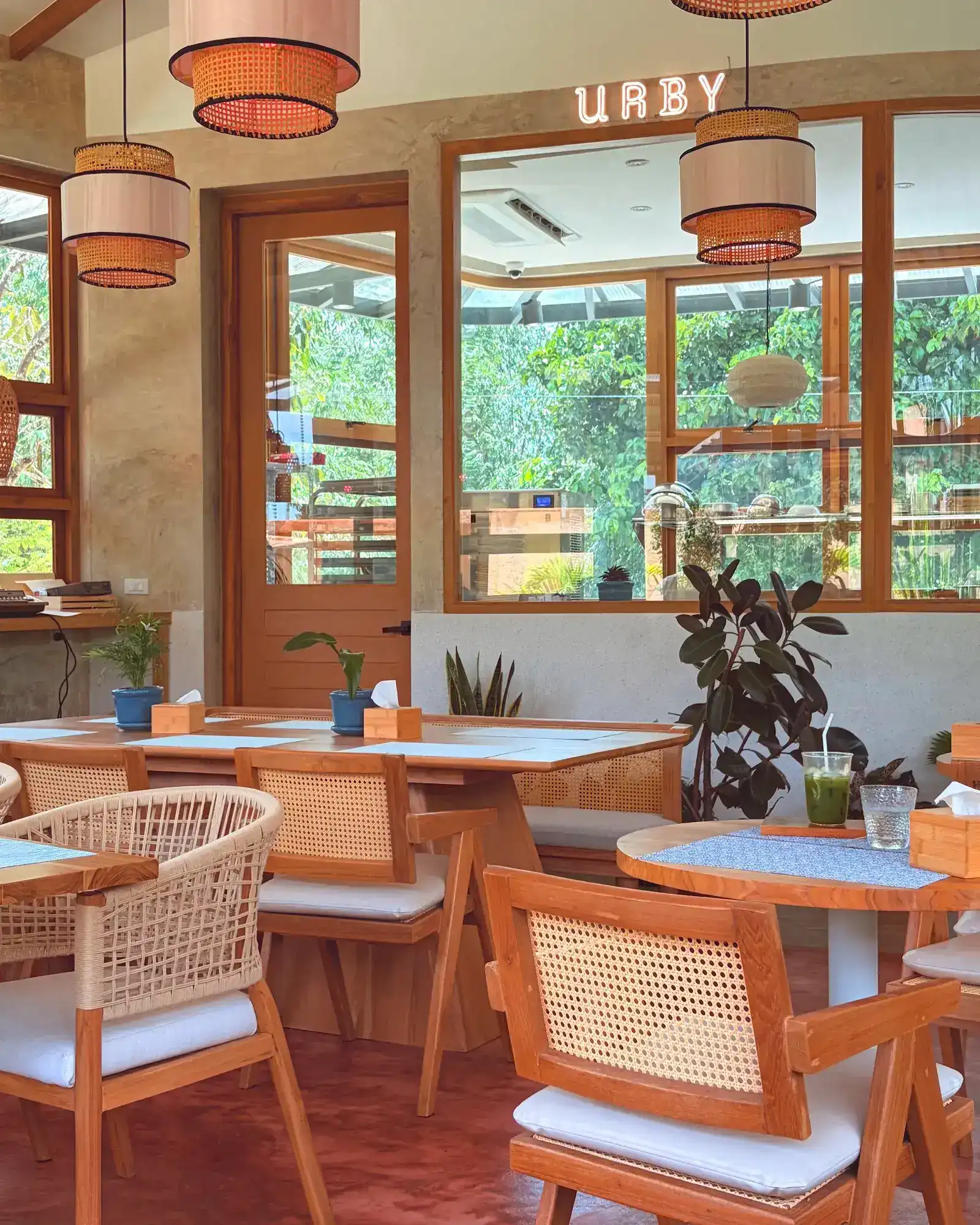 Inside URBY: teak chairs with rattan backs, rattan lamps and teak-framed windows onto the trees