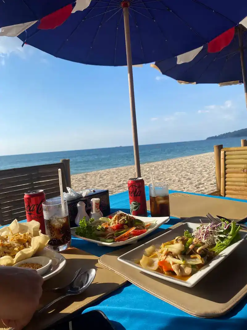 Plates of Thai food on a blue tablecloth under a navy umbrella, the sea behind