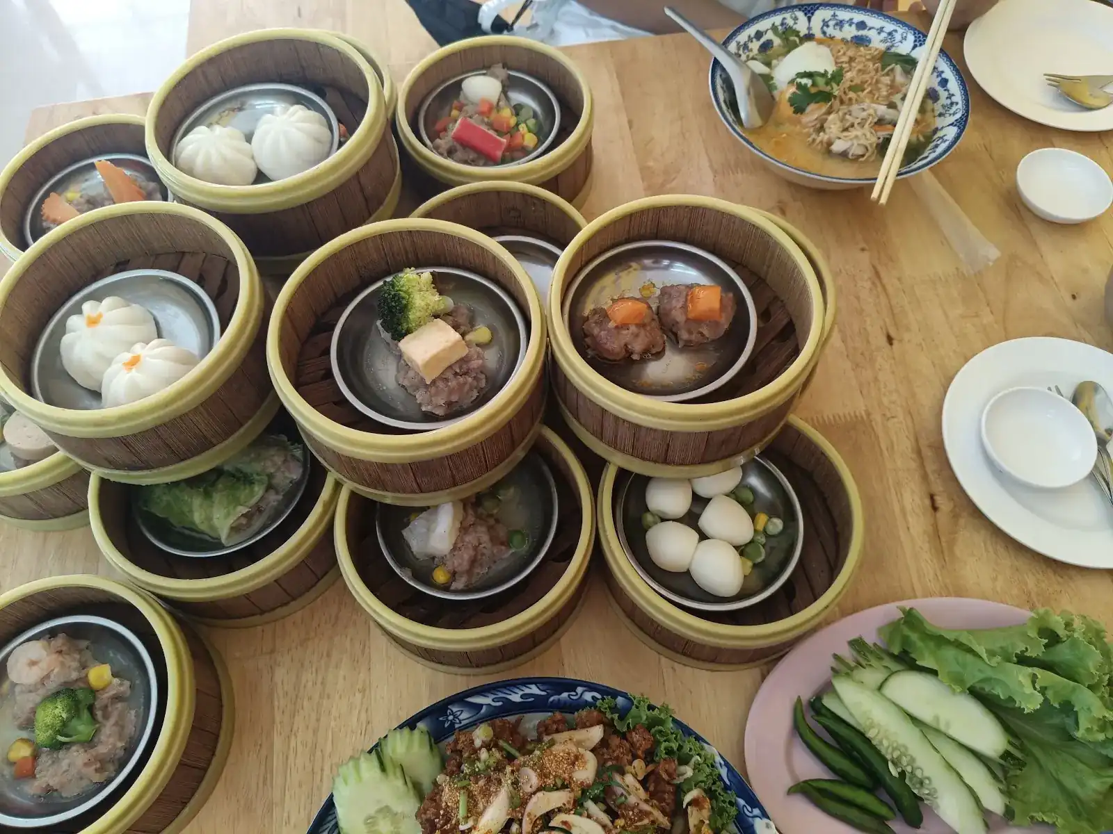Bamboo steamers of dim sum on a wooden table: steamed buns, pork dumplings and cabbage rolls, with a bowl of noodle soup and fresh greens