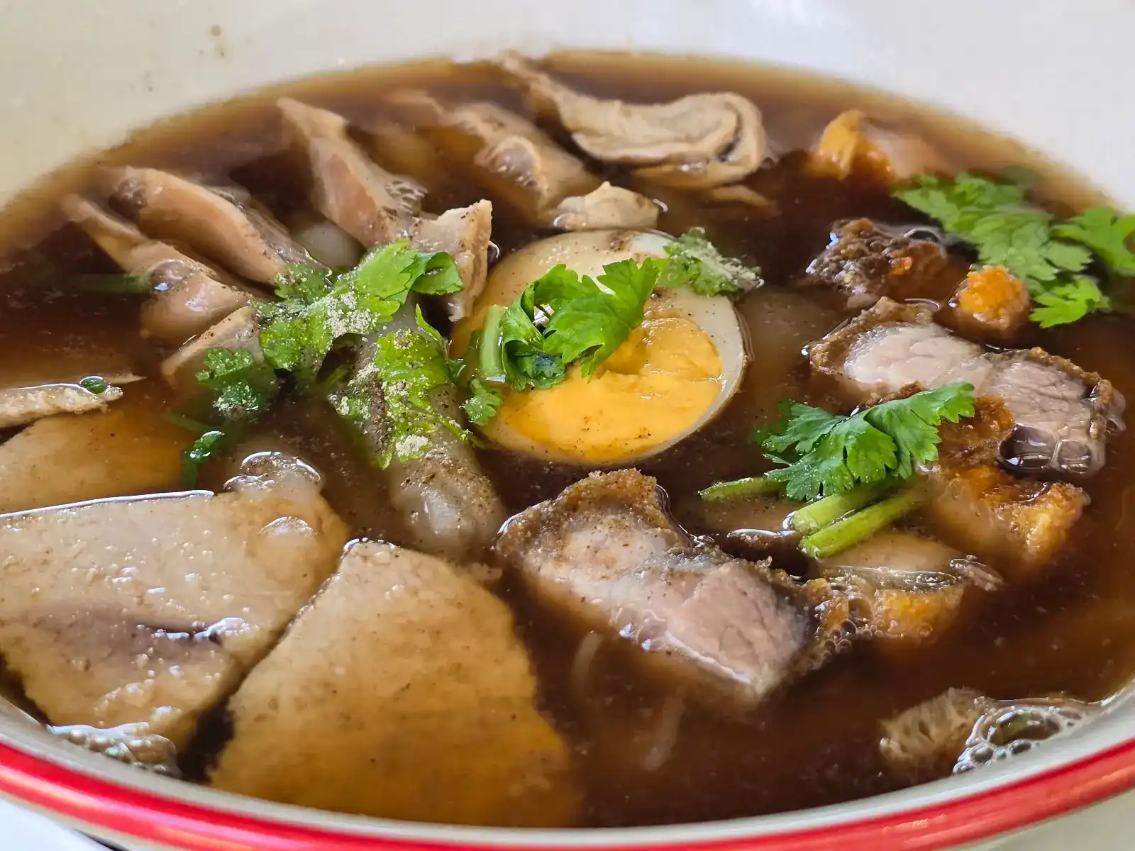 A bowl of rolled noodle soup with crispy pork, sliced pork, a halved egg and coriander in dark broth