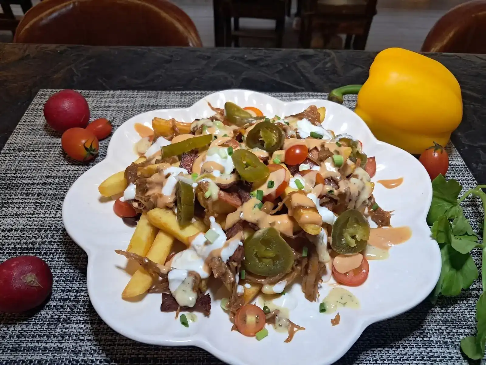 Fries piled with shredded meat, jalapeño slices, cherry tomatoes and sauces on a white plate
