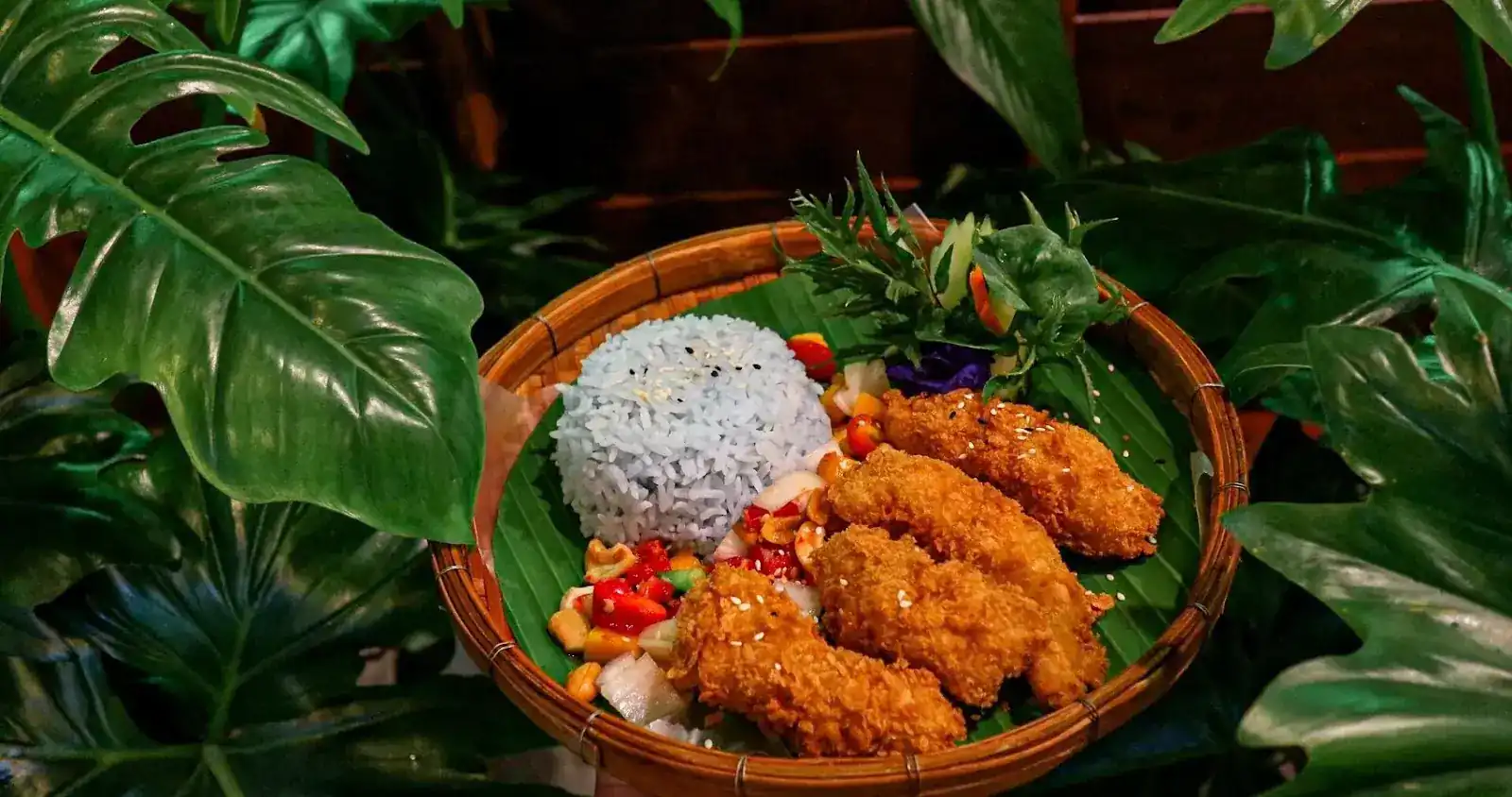 Crumbed fried chicken with blue rice and a chopped salad with cashews, on a banana leaf in a woven basket