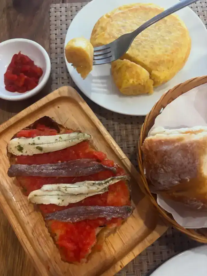 A tosta with white and salted anchovies on tomato, a potato omelette and a basket of bread, seen from above
