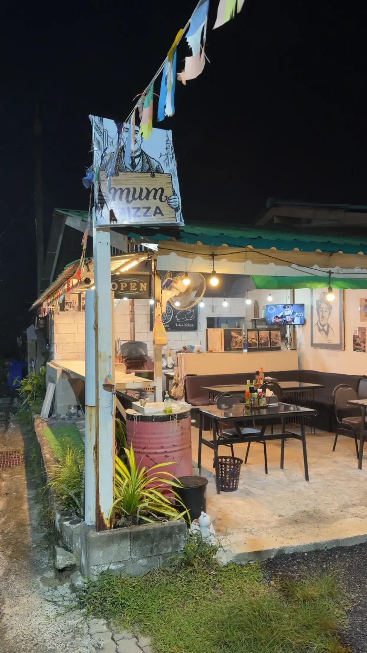 MUM Pizza at night: the bowler-hat sign above an open-air shop with a green roof, string lights and tables