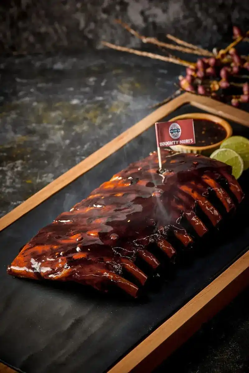 A whole rack of glazed BBQ spare ribs on a wood-framed slate board, with lime and dipping sauce