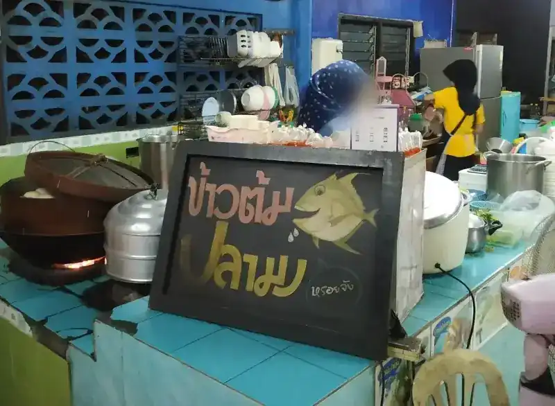 The hand-painted ข้าวต้มปลามง sign with its gold fish, standing on the turquoise-tiled counter
