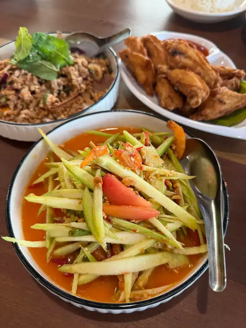 A pounded salad with tomato in a white bowl, with larb and fried chicken wings behind