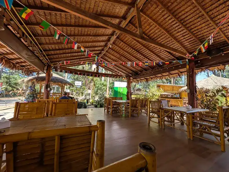A dining pavilion with a thatched ceiling, bamboo tables and strings of flags