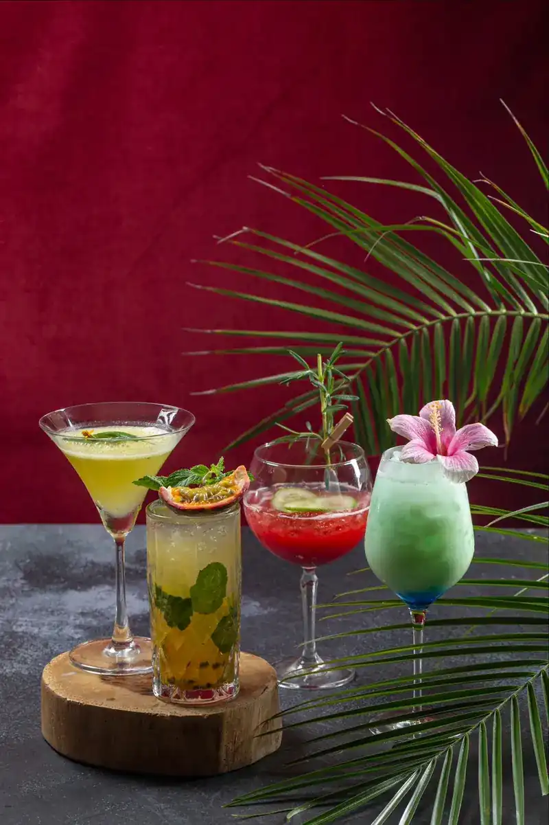 Four colourful drinks with mint, fruit and a flower, against a red wall and palm leaves